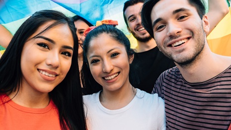 Five friends taking a selfie together at an LGBTQI Pride event. Hispanic and caucasian ethnicities. They are looking at camera and smiling.